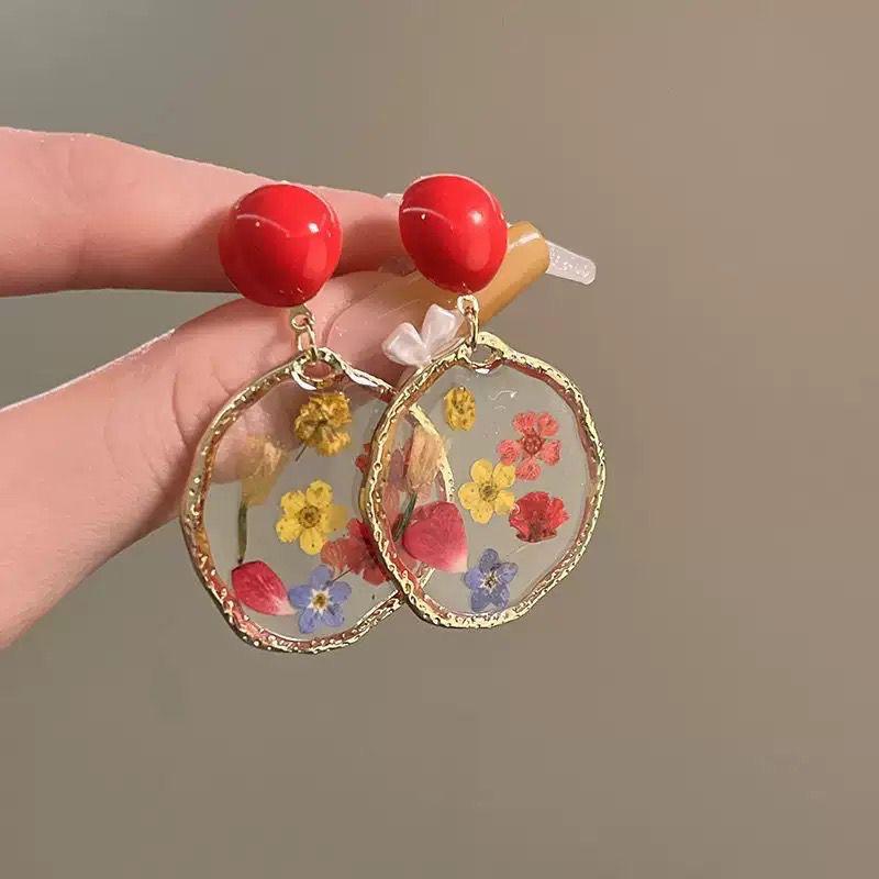 Earrings with red beads and floral designs held by a hand against a neutral background